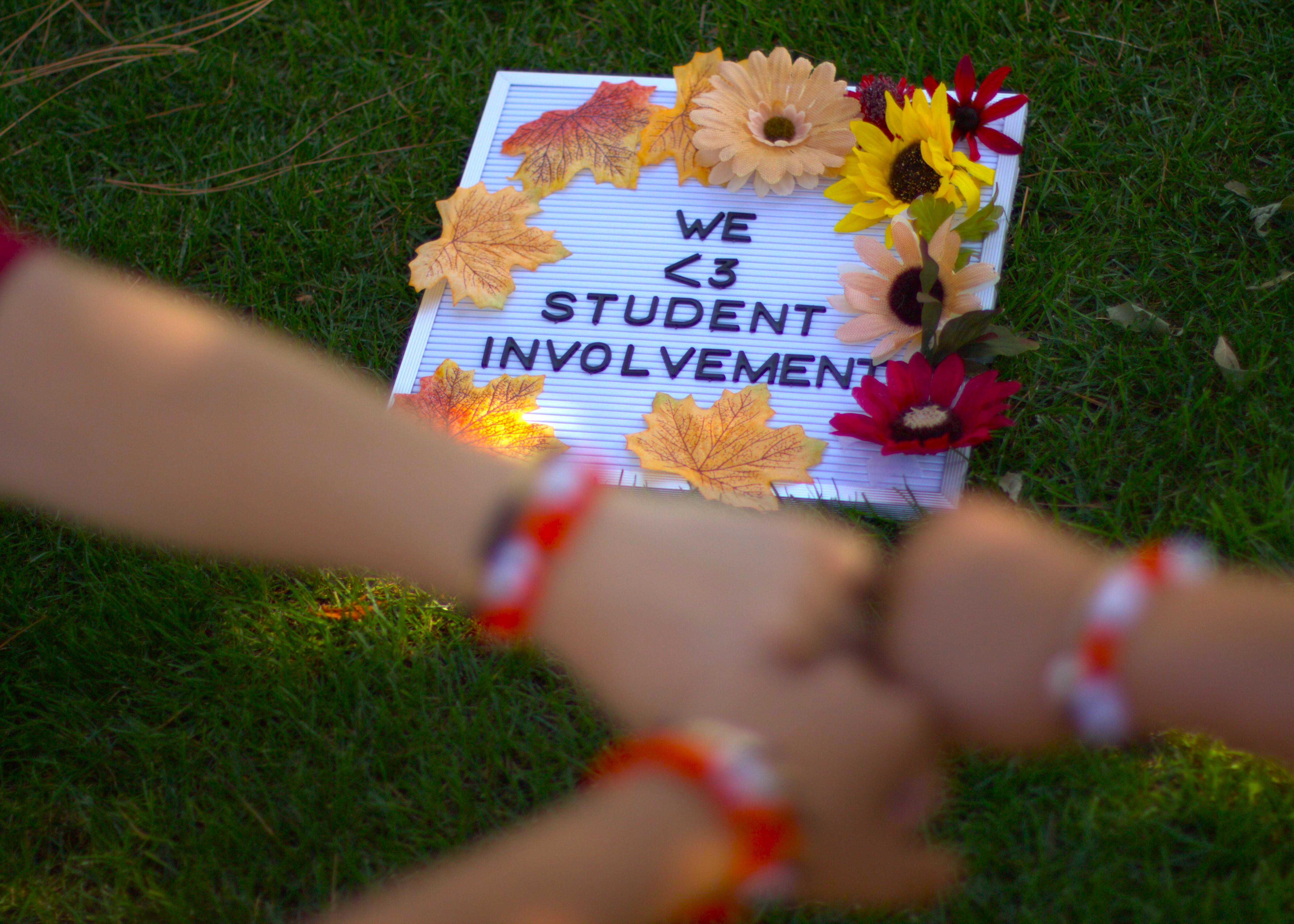 Three out-of-focus hands fist-bump in front of a board that says: "We <3 Student Involvement"